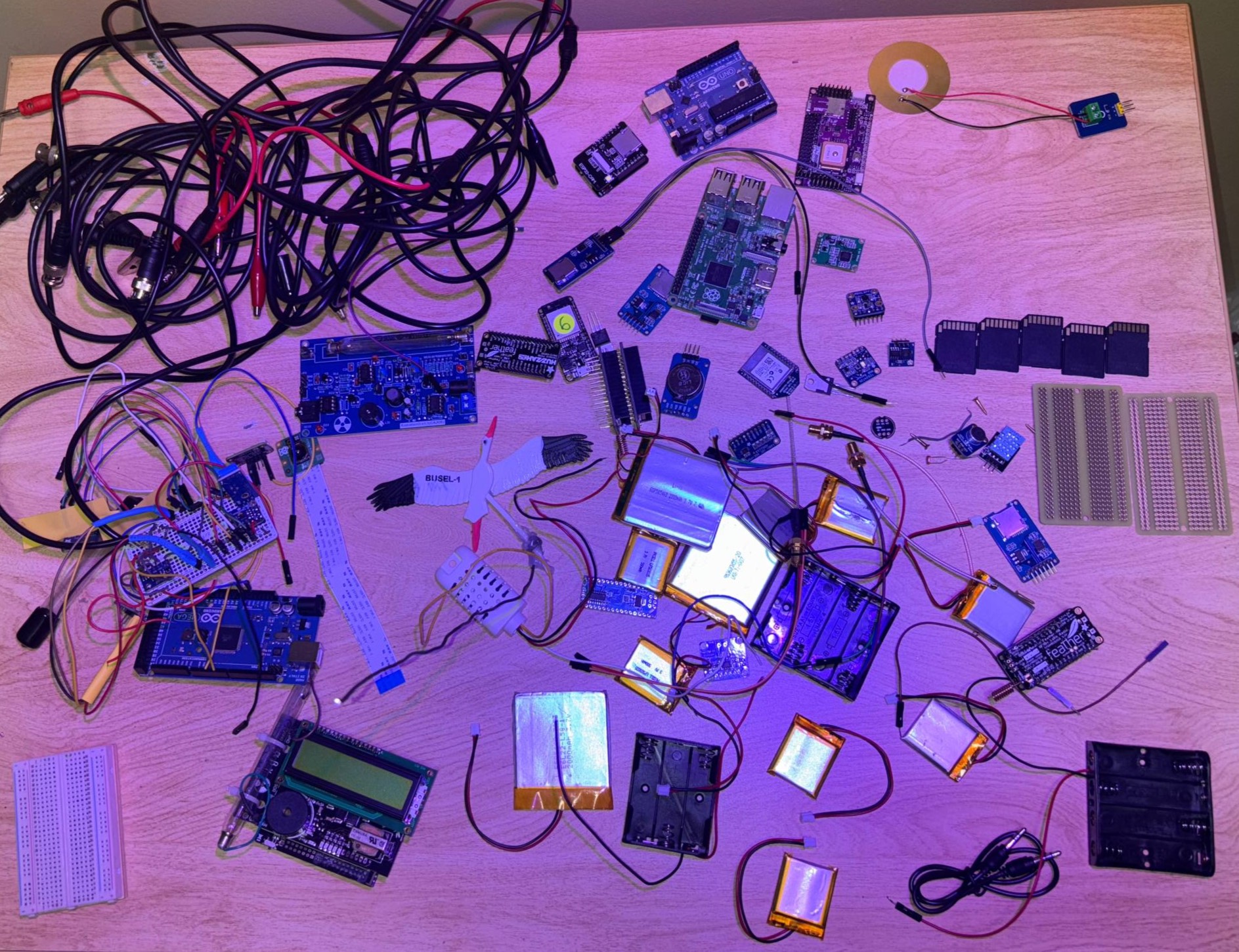 Full electronics inventory laid out on a lab bench: Arduino Mega, Feather M0, sensors, batteries, cables, and wiring spread across the table